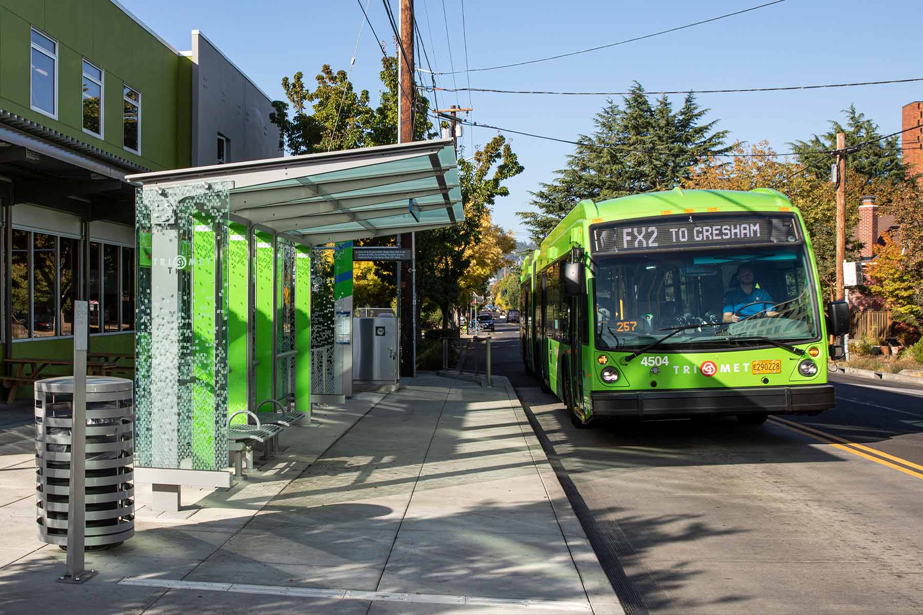 FX bus pulling up to an FX station platform that extends into the street. The station is in front of the sidewalk and includes a glass shelter with weather protection, a digital information sign, benches, and schedule information.